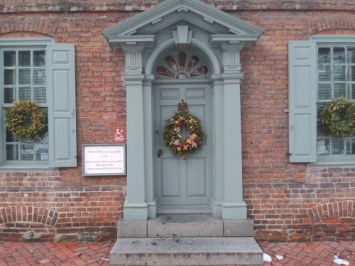 A green door with a decorative wreath, flanked by windows, set against a brick wall with a sign nearby.