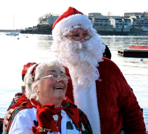 A joyful woman in festive attire smiles at a man dressed as Santa Claus by the water, with buildings in the background.