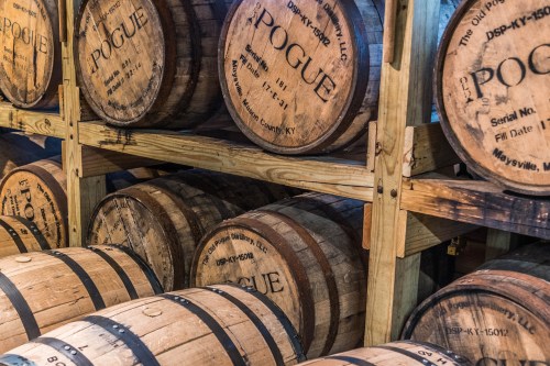 A stack of wooden barrels labeled "Pogue" stored on wooden racks in a distillery.
