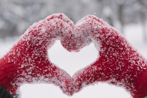 Two red-gloved hands form a heart shape, covered in snow, against a snowy background.