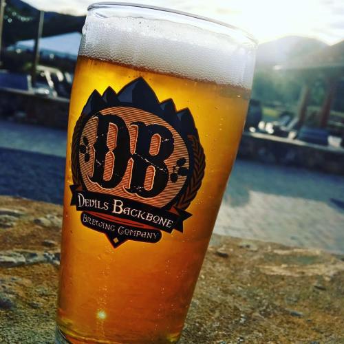 A glass of amber beer with a logo from Devils Backbone Brewing Company, set against a scenic outdoor background.