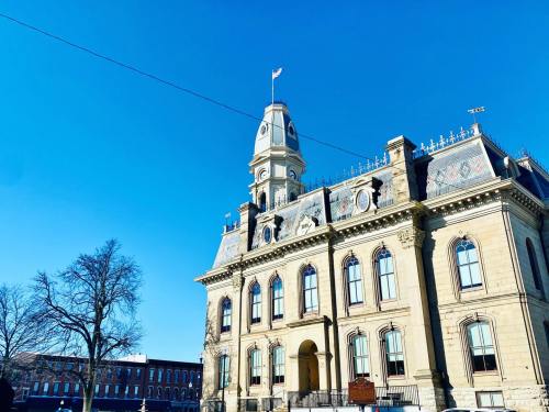 Historic building with a clock tower and flag, set against a clear blue sky. Trees are visible in the foreground.
