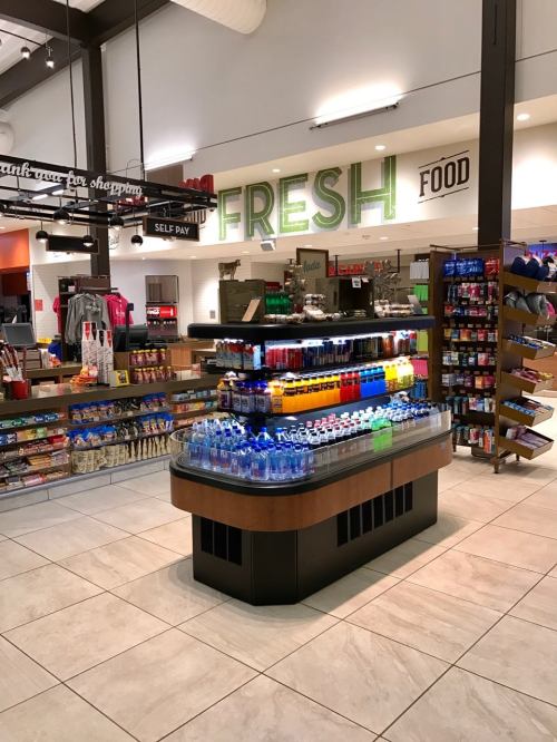 A brightly lit convenience store with a colorful beverage display, snacks, and a "Fresh Food" sign in the background.