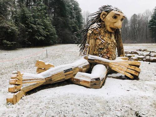 A large wooden sculpture of a troll-like figure sitting in a snowy field, surrounded by trees.