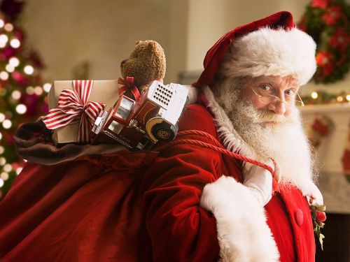 Santa Claus in a red suit smiles while carrying a sack filled with gifts and a teddy bear on his back.