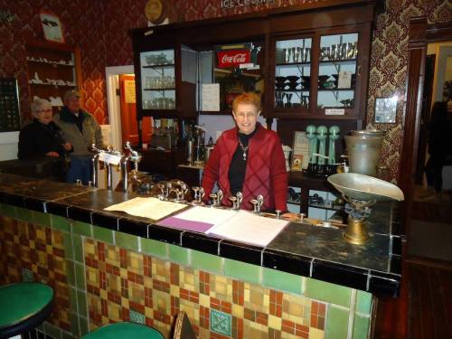 A woman stands behind a vintage bar with taps, surrounded by historical decor and patrons in a cozy setting.