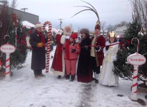 A festive scene with Santa, children, and costumed characters by Christmas trees, celebrating at a winter event.