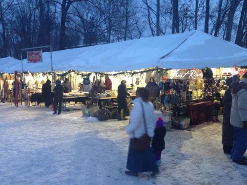 A winter market with tents, lights, and people browsing goods in a snowy landscape. Trees are visible in the background.