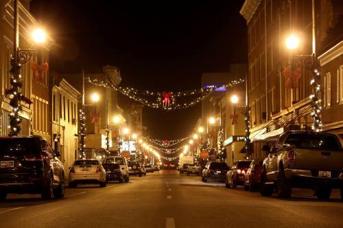 A festive street at night, lined with cars and decorated with holiday lights and ornaments.