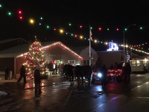 A festive scene at night with a decorated Christmas tree, colorful lights, and a horse-drawn carriage in a snowy setting.