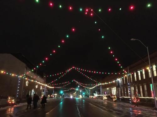 Colorful holiday lights strung across a street at night, with people walking and festive decorations lining the buildings.