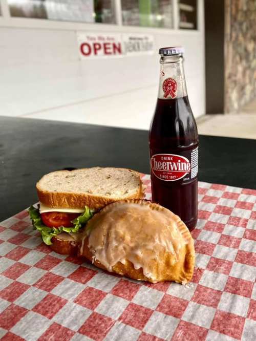 A sandwich with lettuce and tomato beside a glazed pastry and a bottle of Cheerwine on a checkered paper.