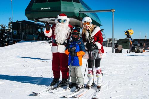 Santa, a child in ski gear, and a woman in festive attire pose together on a snowy ski slope with a ski lift in the background.