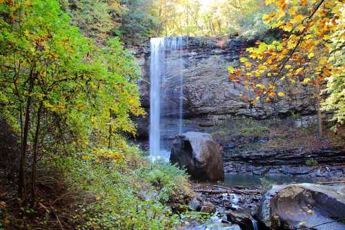 A serene waterfall cascades down rocky cliffs, surrounded by vibrant autumn foliage and a tranquil pool below.