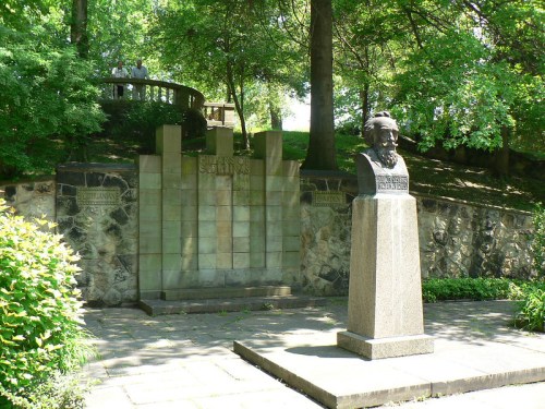 A stone monument with a bust stands in a green park, surrounded by trees and a stone wall.