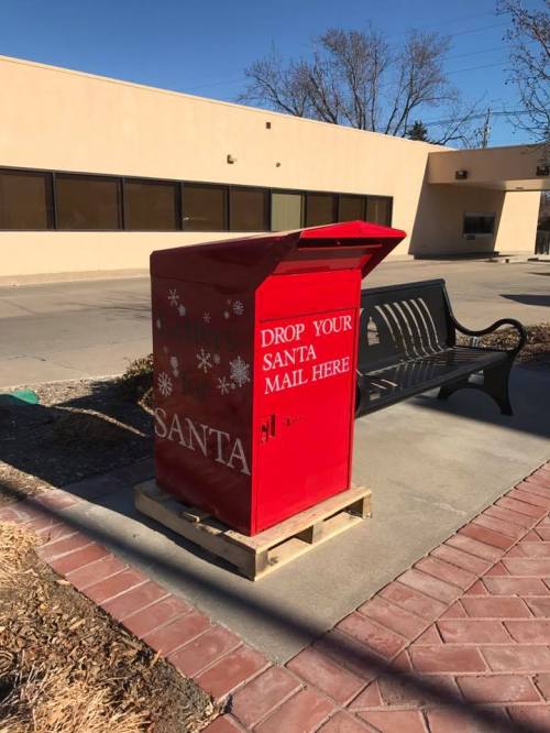 A bright red mailbox labeled "DROP YOUR SANTA MAIL HERE" sits on a sidewalk near a bench.