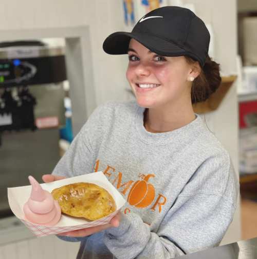 A smiling young woman in a gray sweatshirt and black cap holds a dessert plate with a pastry and soft serve ice cream.