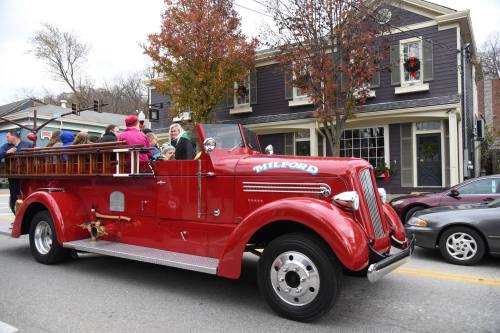 A vintage red fire truck drives past a festive building with holiday decorations and people enjoying the ride.