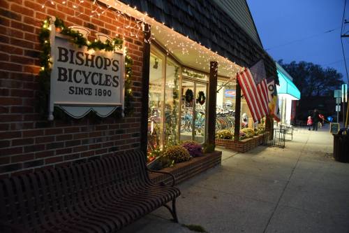 A bicycle shop with festive lights, an American flag, and a bench outside, set in a cozy evening atmosphere.