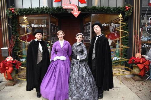 Four people in Victorian costumes stand outside a festive shop, decorated for the holidays with wreaths and lights.