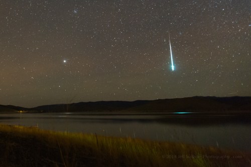 A night sky filled with stars, featuring a bright meteor streaking across above a calm lake.