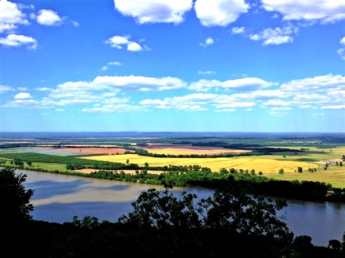 A panoramic view of lush fields and a river under a bright blue sky with fluffy clouds.