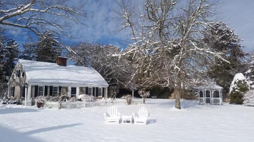 A snow-covered house with white chairs in the foreground and a gazebo in a winter landscape.