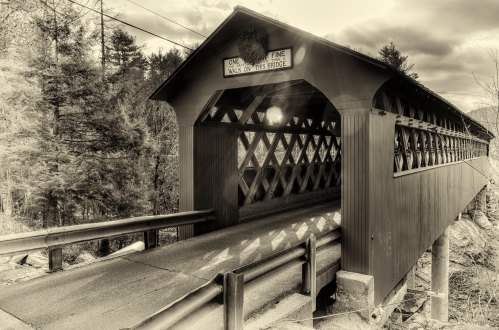 A black-and-white photo of a covered bridge surrounded by trees, with a sign reading "One Fine Walk on This Bridge."
