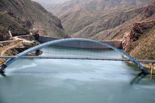 A blue arch bridge spans a calm river, surrounded by rugged mountains and a dam in the background.