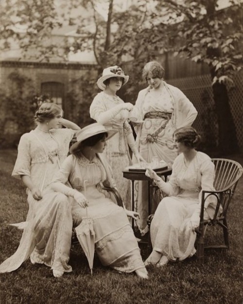 Five women in vintage dresses and hats gather in a garden, engaged in conversation around a small table.