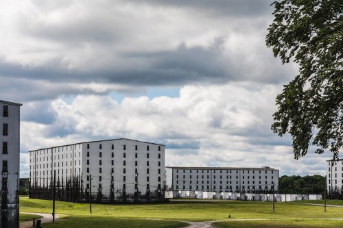 Abandoned buildings with white exteriors and darkened lower sections, set against a cloudy sky and green grass.