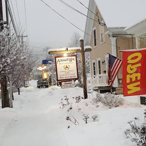 Snow-covered street scene featuring a restaurant sign, American flag, and "Open" banner amidst winter weather.