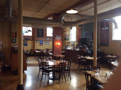 Cozy restaurant interior with wooden tables, chairs, and colorful artwork on the walls. Soft lighting creates a warm atmosphere.