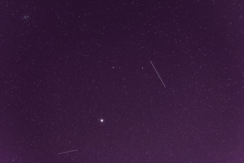 A starry night sky filled with numerous stars, featuring a few bright celestial objects and streaks of light.