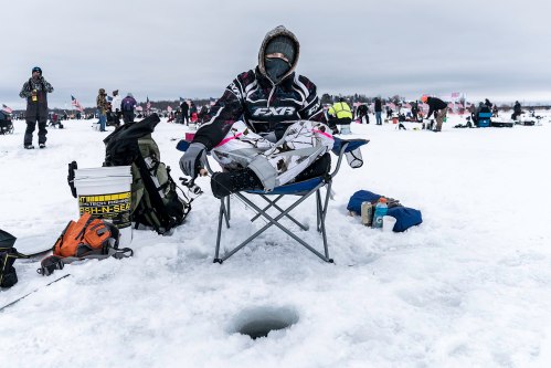 A person in winter gear sits on a chair by an ice fishing hole, surrounded by others on a snowy landscape.