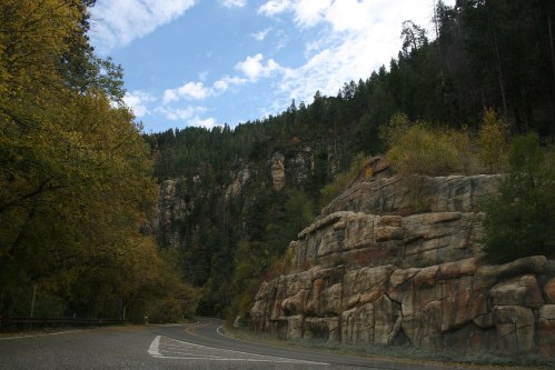 A winding road surrounded by tall trees and rocky cliffs under a partly cloudy sky.