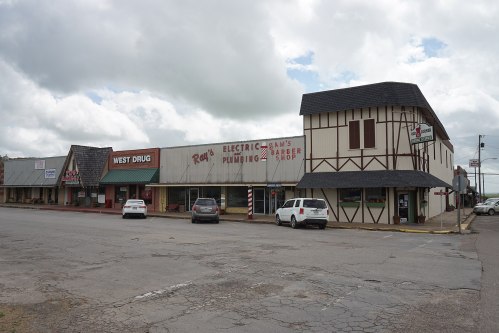 A small town street featuring a row of shops, including a drugstore and a barber shop, under a cloudy sky.