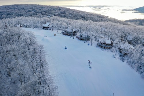 A snowy landscape with frosted trees and cabins, overlooking a misty valley under a cloudy sky.