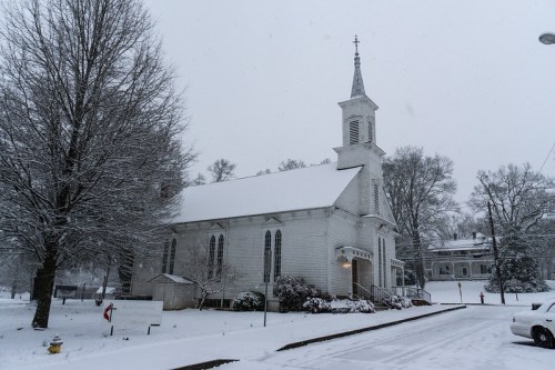 A snow-covered church with a tall steeple, surrounded by trees and a quiet street in a winter landscape.