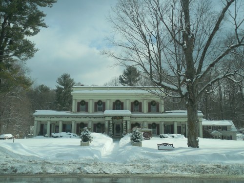 A large, elegant house with white columns, surrounded by snow-covered trees and a snowy yard on a cloudy day.