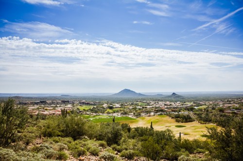 A scenic view of a golf course surrounded by desert landscape and mountains under a blue sky with scattered clouds.
