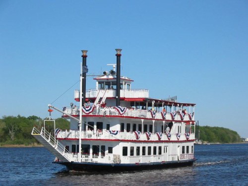 A decorated riverboat with multiple decks sails on a calm river under a clear blue sky.