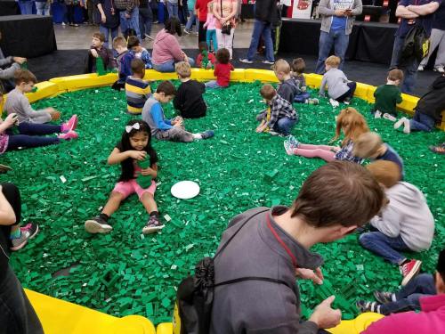 A large group of children playing in a circular area filled with green building blocks, with adults nearby.