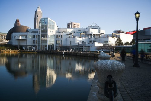 A modern building reflects in calm water, with a city skyline and a lamppost in the foreground.