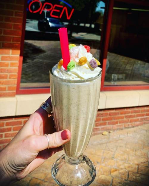 A hand holds a tall milkshake topped with whipped cream and colorful candy, with an "OPEN" sign in the background.