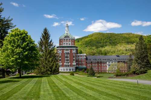 A grand brick building with a tower, surrounded by lush green trees and a hillside under a clear blue sky.