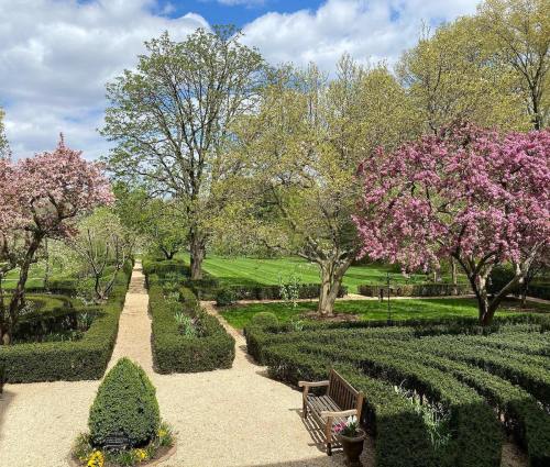 A serene garden with neatly trimmed hedges, blooming pink trees, and a gravel path under a partly cloudy sky.