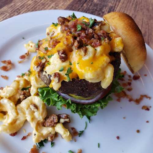 A gourmet burger topped with macaroni and cheese, bacon, and cheddar, served with a side of macaroni on a white plate.