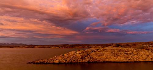 A serene landscape featuring a rocky shoreline under a vibrant sunset with colorful clouds reflecting in the water.