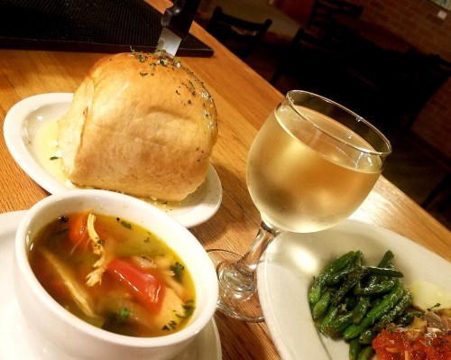 A bowl of soup with vegetables, a roll with butter, a glass of white wine, and a plate of green beans on a wooden table.